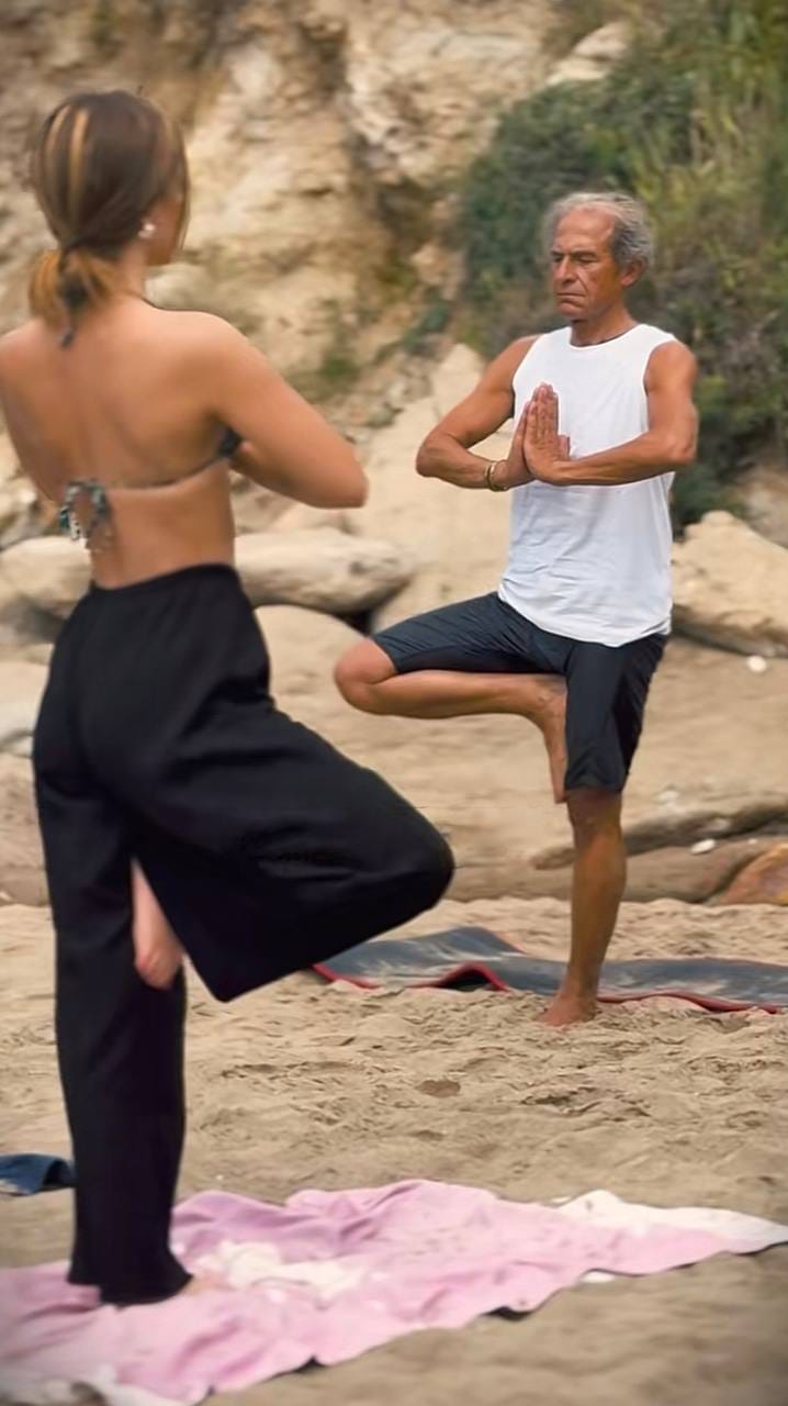 Mario Funari insegna yoga sulla spiaggia, nella posizione dell'albero, con un'allieva di spalle in primo piano Mario Funari insegna yoga sulla spiaggia, nella posizione dell'albero, con un'allieva di spalle in primo piano